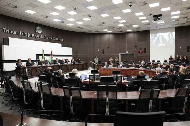 Sala de tribunal do Tribunal de Justiça de Santa Catarina. A sala é ampla, com um teto repleto de painéis de iluminação. Há várias pessoas sentadas em cadeiras ao redor de uma grande mesa em formato de "U". Na parede ao fundo, está escrito "Tribunal de Justiça de Santa Catarina". Há uma tela de projeção mostrando uma imagem de uma pessoa falando. Algumas bandeiras estão posicionadas ao lado esquerdo da sala. Várias pessoas estão vestidas formalmente, possivelmente juízes e advogados. Há também algumas pessoas em pé ao fundo, incluindo seguranças.