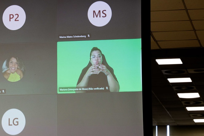 Tela de uma videoconferência com várias janelas de participantes. No canto superior esquerdo, há um círculo com as iniciais "P2". Ao lado, outro círculo com as iniciais "MS" e o nome "Marina Mabor Schelemberg" abaixo. No centro, uma janela maior mostra uma mulher com cabelo preso e óculos, fazendo sinais em Libras, com um fundo verde. A legenda abaixo dela diz "Mariana (intérprete de libras) (Não verificado)". No canto inferior esquerdo, há um círculo com as iniciais "LG". Ao lado, uma janela com a imagem de uma mulher sorrindo, vestindo uma blusa amarela. À direita, parte do teto de uma sala com luminárias visíveis.