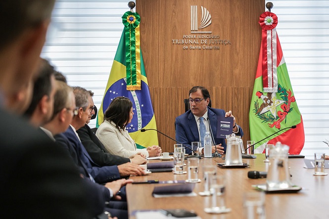 Reunião em uma sala de conferências com várias pessoas sentadas ao redor de uma mesa de madeira. No centro, um homem de terno azul está segurando um documento. Ao fundo, há duas bandeiras, uma do Brasil e outra do estado de Santa Catarina. Na parede, está escrito "Poder Judiciário Tribunal de Justiça de Santa Catarina". As pessoas ao redor da mesa estão vestidas formalmente, e há copos de água e garrafas sobre a mesa. A atmosfera parece ser de uma reunião oficial ou cerimônia.