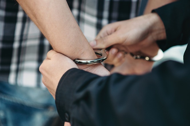 Duas pessoas estão na imagem. Uma delas está colocando algemas nos pulsos da outra. A pessoa que está sendo algemada veste uma camisa xadrez, enquanto a outra pessoa veste uma camisa preta. A cena sugere uma situação de prisão ou detenção.