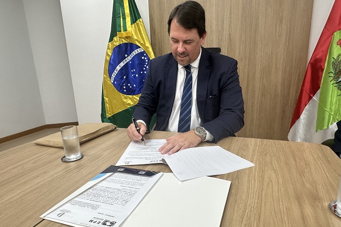 Um homem de terno azul escuro e gravata listrada está sentado em uma mesa de madeira, assinando documentos. Ele tem cabelo castanho escuro e usa um relógio no pulso esquerdo. À sua frente, há um copo de água sobre a mesa. Atrás dele, estão as bandeiras do Brasil e de um estado brasileiro, possivelmente Santa Catarina, devido à presença de uma bandeira com um brasão que lembra o do estado. A sala tem paredes brancas e um painel de madeira ao fundo.