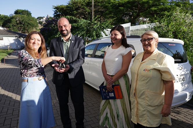 Quatro pessoas estão em pé em frente a um carro branco. À esquerda, uma mulher de cabelos castanhos e longos, vestindo uma blusa estampada e saia azul clara, está entregando uma chave para um homem careca, de barba, usando um terno escuro. Ao lado dele, uma mulher de cabelos castanhos e lisos, vestindo uma blusa branca e saia listrada em verde e branco, segura um livro azul e laranja. À direita, uma mulher de óculos, cabelos curtos e grisalhos, veste um uniforme bege com o logotipo "APAE" bordado. Ao fundo, há árvores e uma cerca branca. O clima parece ensolarado.