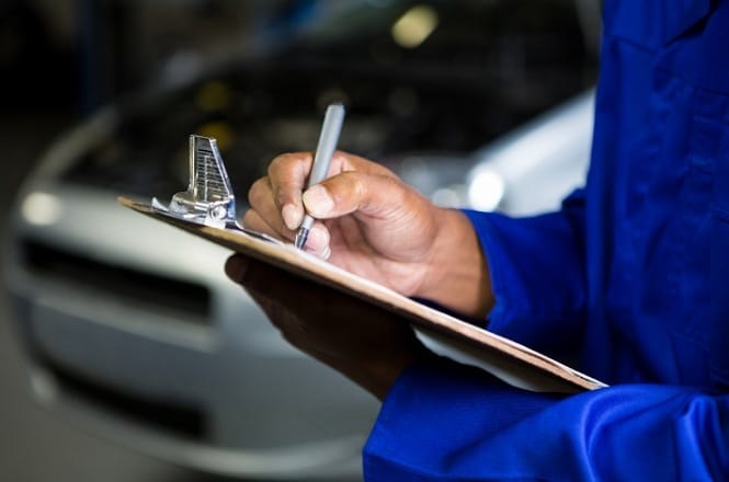 Uma pessoa vestindo um macacão azul está segurando uma prancheta e escrevendo nela com uma caneta. Ao fundo, há um carro desfocado, sugerindo que a cena ocorre em uma oficina mecânica ou local de manutenção de veículos. A atenção está focada nas mãos da pessoa e na prancheta.
