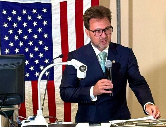 Homem de terno azul escuro e gravata verde claro, segurando um microfone em uma mão e gesticulando com a outra, como se estivesse falando em público. Ele está em pé atrás de uma mesa que tem um monitor de computador e um dispositivo de câmera com a inscrição "12X". Ao fundo, há uma bandeira dos Estados Unidos com listras vermelhas e brancas e um campo azul com estrelas brancas. O homem usa óculos e tem cabelo castanho.