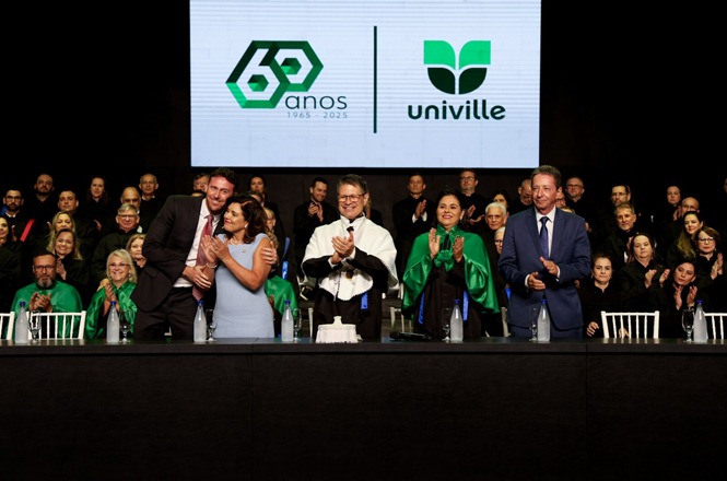 Um grupo de pessoas está em um palco, aparentemente durante uma cerimônia ou evento formal. No centro, há uma mesa comprida com várias garrafas de água e copos. Atrás da mesa, cinco pessoas estão em pé, aplaudindo. A pessoa no centro veste uma beca preta e branca, enquanto as outras usam trajes formais, com alguns usando capas verdes. Ao fundo, há mais pessoas sentadas, também em trajes formais. Na parede atrás do grupo, há um grande painel com o logotipo e o nome "univille", além de um emblema que diz "60 anos 1965 - 2025".