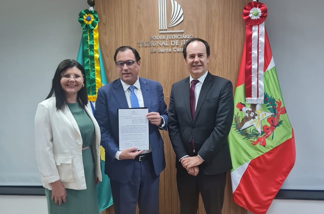 Três pessoas estão posando para uma foto em frente a uma parede de madeira com o logotipo do "Poder Judiciário Tribunal de Justiça de Santa Catarina". À esquerda, uma mulher de cabelos escuros usa óculos e veste um blazer branco sobre um vestido verde. No centro, um homem de óculos, vestindo um terno azul e gravata azul clara, segura um documento. À direita, outro homem veste um terno preto com gravata vermelha. Atrás deles, há duas bandeiras: a do Brasil à esquerda e a de Santa Catarina à direita.
