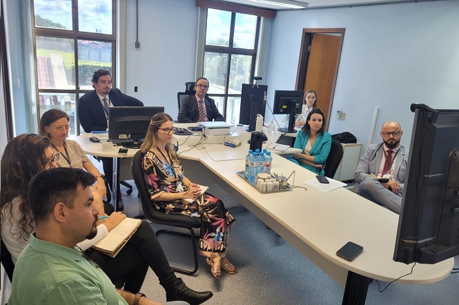 Sala de reunião com nove pessoas sentadas ao redor de uma mesa em formato de "U". Na mesa, há computadores, garrafas de água e papéis. As pessoas estão vestidas formalmente, algumas com ternos e outras com roupas sociais. A sala tem janelas grandes ao fundo, permitindo a entrada de luz natural. Todos estão voltados para uma tela grande, possivelmente assistindo a uma apresentação ou conferência.