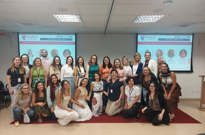 Um grupo de aproximadamente 25 pessoas, composto principalmente por mulheres, está posando para uma foto em um ambiente interno, provavelmente uma sala de conferências ou auditório. Algumas pessoas estão de pé, enquanto outras estão agachadas ou sentadas na frente. Ao fundo, há duas telas de projeção que exibem informações sobre um evento, mencionando "MARCO" e "19 MAR", além de um canal no YouTube chamado "CANAL TSC". As pessoas na foto estão sorrindo e parecem estar participando de um evento ou reunião. O ambiente é bem iluminado, com teto de placas brancas e luminárias embutidas.