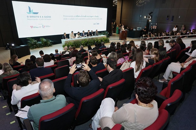 Auditório com várias pessoas sentadas em cadeiras vermelhas, assistindo a um evento. Na frente, há uma mesa com sete pessoas sentadas, provavelmente palestrantes ou autoridades. Atrás deles, um grande telão exibe a mensagem: "O DIREITO E A SAÚDE Respeitando a Judicialização. Promovendo um direito à saúde mais seguro e ágil, onde o cuidado acontece de forma rápida e eficiente." O ambiente é bem iluminado e há plantas decorativas em frente à mesa dos palestrantes. Algumas pessoas estão tirando fotos ou gravando o evento com seus celulares.