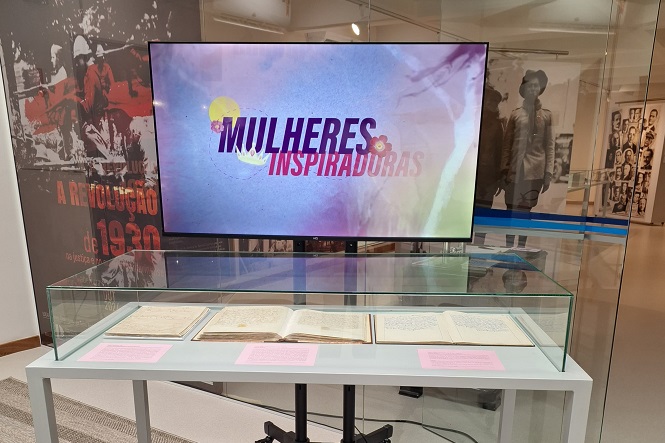 Em um ambiente de exposição, há uma televisão exibindo a frase "MULHERES INSPIRADORAS" em letras grandes. Abaixo da televisão, há uma vitrine de vidro contendo livros ou documentos abertos, com etiquetas rosa em frente a cada um. Ao fundo, há um painel com a imagem de pessoas e o texto "A REVOLUÇÃO de 1930". O ambiente parece ser um museu ou uma galeria, com outras imagens e exibições visíveis ao fundo.