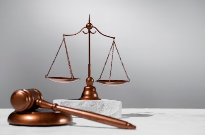 Um martelo de juiz de madeira está em primeiro plano, repousando sobre uma base circular. Ao fundo, há uma balança de justiça de cor bronze, com dois pratos suspensos por correntes. A balança está sobre uma superfície clara e o fundo é de cor cinza suave. A imagem transmite uma sensação de justiça e legalidade.