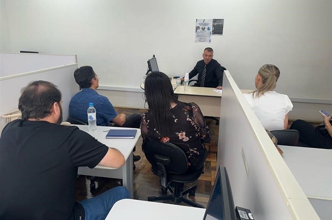 Sala de escritório com seis pessoas. Cinco delas estão sentadas em cadeiras de escritório em torno de uma mesa, enquanto um homem de terno está sentado do outro lado da mesa, parecendo conduzir uma reunião ou apresentação. As pessoas estão prestando atenção ao homem de terno. Há divisórias brancas entre as mesas, e uma garrafa de água está sobre uma das mesas. Na parede ao fundo, há dois cartazes. O ambiente parece ser um escritório ou sala de reuniões.