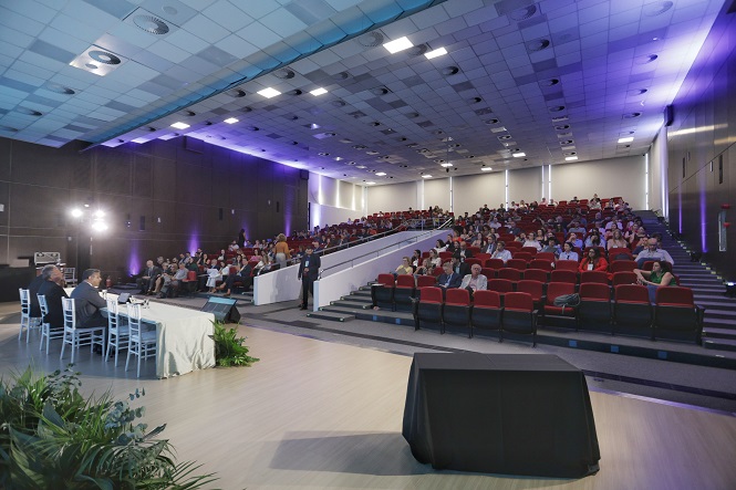 Auditório amplo com cadeiras vermelhas, parcialmente ocupado por pessoas. À esquerda, há uma mesa coberta com uma toalha branca, onde estão sentadas quatro pessoas, aparentemente participando de um painel ou conferência. O teto é alto, com iluminação embutida, e as paredes são escuras, com iluminação azul suave em alguns pontos. Há plantas decorativas na frente da mesa. O ambiente parece formal e bem organizado.