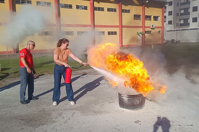 Uma mulher está usando um extintor de incêndio para apagar chamas que estão saindo de um recipiente cilíndrico no chão. Ao lado dela, um homem observa a ação. Ambos estão em um espaço aberto, com um prédio de paredes amarelas e detalhes vermelhos ao fundo. O homem está vestindo uma camiseta vermelha e calças escuras, enquanto a mulher usa uma blusa marrom e calças jeans. Há fumaça ao redor das chamas e uma sombra projetada no chão.