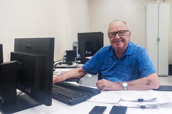 Homem idoso sentado em uma mesa de escritório, sorrindo para a câmera. Ele usa óculos e uma camisa polo azul. Na mesa, há dois monitores de computador, um teclado, papéis e dois celulares. Ao fundo, há um armário branco e mais equipamentos de escritório. O ambiente parece ser um escritório moderno e bem iluminado.