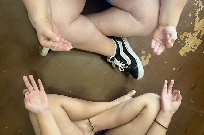 Duas pessoas estão sentadas no chão em posição de meditação, formando um círculo. Ambas têm as pernas cruzadas e as mãos repousando sobre os joelhos, com as palmas voltadas para cima e os dedos em posição de mudra. Uma das pessoas está usando um tênis preto com detalhes brancos, enquanto a outra está descalça e usa uma tornozeleira. O chão parece ser de madeira, com algumas áreas desgastadas.