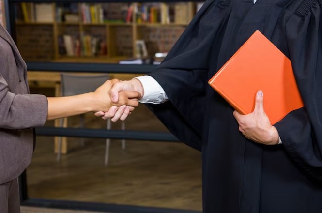 A imagem mostra duas pessoas apertando as mãos. Uma delas veste uma toga preta e segura um livro ou pasta laranja na outra mão. O fundo parece ser um ambiente interno com prateleiras de livros.
