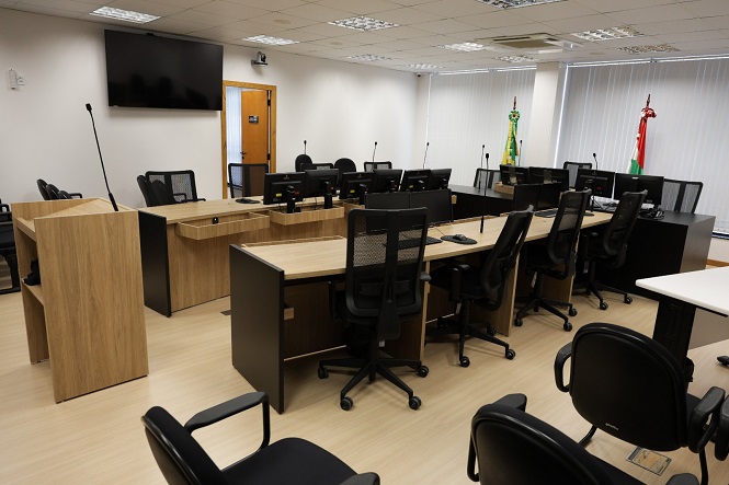 A imagem mostra uma sala de tribunal vazia. Há várias mesas de madeira, equipadas com microfones e cadeiras giratórias. Na parte frontal, há dois púlpitos para advogados e testemunhas. No fundo, encontram-se bandeiras e uma grande televisão na parede. A iluminação é fornecida por lâmpadas no teto e a sala possui janelas com persianas.