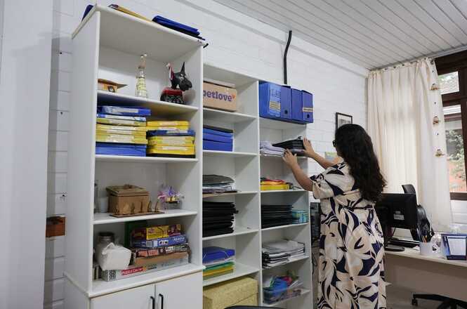 A imagem mostra uma pessoa organizando prateleiras em um ambiente interno, provavelmente um escritório ou espaço de trabalho. Há várias prateleiras brancas cheias de documentos, pastas e alguns objetos decorativos. A pessoa está de pé ao lado das prateleiras, com as mãos levantadas, como se estivesse rearranjando ou pegando algo. À direita, há uma mesa com um computador, e à esquerda, mais prateleiras com materiais diversos. As paredes são brancas e há uma janela com cortinas ao fundo.