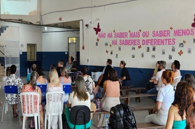 Na imagem, há um grupo de pessoas sentadas, assistindo a uma apresentação ou reunião em um ambiente que parece ser uma escola ou centro comunitário. As cadeiras são de diferentes tipos, algumas de plástico e outras de madeira. Na parede, há uma decoração com borboletas e uma frase que diz: "NÃO HÁ SABER MAIS OU SABER MENOS, HÁ SABERES DIFERENTES". A sala tem um aspecto simples, com paredes pintadas em dois tons, branco e azul.