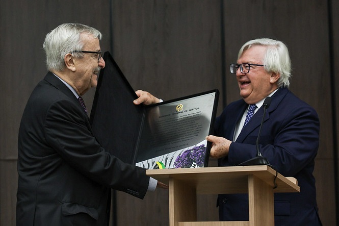Foto horizontal: autoridades trocam placa de homenagem em cerimônia, gesto sorridente, púlpito ao lado, ambiente institucional.
