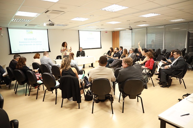 A imagem mostra uma sala de conferência onde várias pessoas estão reunidas. Elas estão sentadas em cadeiras dispostas em semicírculo, prestando atenção em duas pessoas na frente que parecem liderar a reunião. Na tela projetada na parede, o título parece indicar que é uma reunião de assessores. A sala é iluminada por luzes do teto, e há mesas brancas na frente. Algumas pessoas estão fazendo anotações ou levantando as mãos, indicando que é uma sessão interativa.