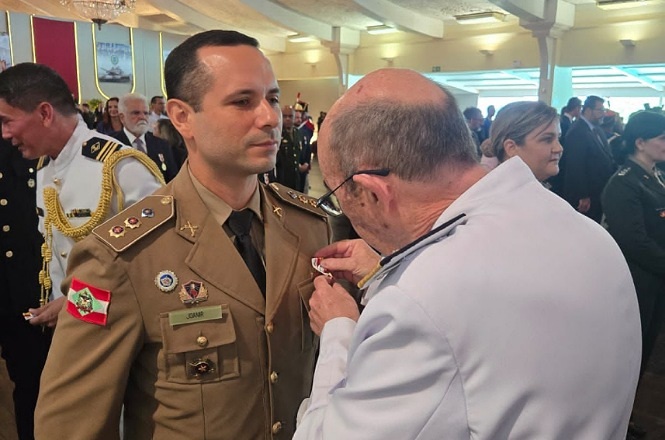 A imagem mostra duas pessoas em um evento formal. A pessoa à esquerda está vestindo um uniforme militar, com várias insígnias e medalhas, e está recebendo algum tipo de condecoração de outra pessoa vestida de branco. No fundo, há outras pessoas em trajes formais, algumas também em uniformes militares, em um ambiente que parece ser um salão de eventos bem iluminado. A atmosfera sugere uma cerimônia oficial ou uma solenidade militar.