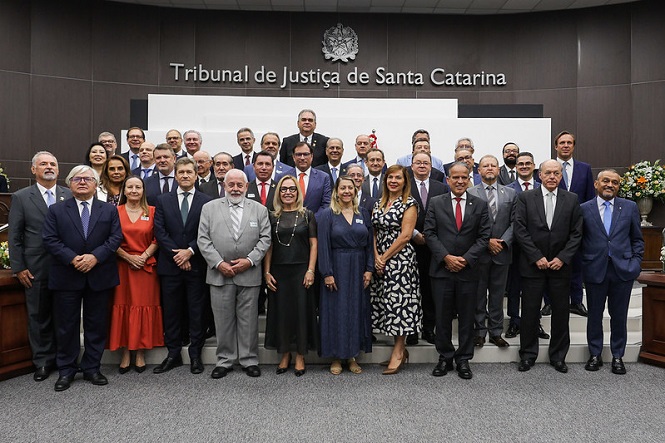 A imagem mostra um grupo de pessoas posando para uma foto em um ambiente formal. Eles estão em pé, bem arrumados, muitos usando ternos e roupas formais. Ao fundo, há um painel com a inscrição "Tribunal de Justiça de Santa Catarina", indicando que o local é uma sala ou auditório relacionado ao tribunal. O ambiente parece ser amplo e bem iluminado, com um piso acarpetado. As pessoas estão organizadas em várias fileiras, sugerindo que se trata de um evento oficial ou reunião importante.