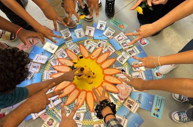 A imagem mostra um grupo de pessoas reunidas em torno de um display colorido no chão. No centro, há um objeto em forma de girassol com pétalas laranja ao redor de um círculo amarelo. Ao redor do girassol, há várias cartas ou rótulos com palavras como "diálogo", "ética", "respeito" e outras, aparentemente em um contexto educativo ou de relacionamento interpessoal. As pessoas estão estendendo as mãos em direção ao centro, algumas fazendo sinais de paz. A cena transmite uma sensação de união e colaboração.