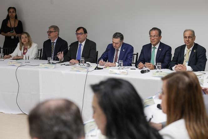 Na imagem, há um grupo de pessoas sentadas em uma mesa comprida coberta com toalhas brancas. Parece ser uma reunião formal ou conferência. Entre os participantes, há homens e mulheres em trajes formais. Alguns deles estão escrevendo ou tomando notas. Há microfones e copos de água na mesa. Ao fundo, uma pessoa está de pé, possivelmente assistindo ou aguardando para falar. O ambiente parece ser um espaço fechado, como uma sala de conferências.