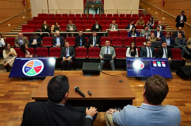 A imagem mostra uma sala de conferências com assentos vermelhos, onde várias pessoas estão sentadas assistindo a uma apresentação. Há duas telas grandes na frente mostrando gráficos. Duas pessoas estão de costas, aparentemente conduzindo a apresentação. O ambiente é formal, sugerindo uma reunião profissional ou empresarial.