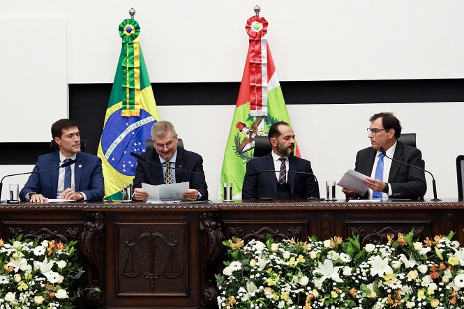 Na imagem, há quatro homens sentados atrás de uma mesa decorada com flores. Atrás deles, estão as bandeiras do Brasil e de outro estado, possivelmente Santa Catarina, devido à sua semelhança com a bandeira dessa unidade federativa. Todos estão vestindo ternos e parecem estar em uma cerimônia ou evento oficial. A mesa apresenta um brasão e a decoração floral é composta por flores brancas e amarelas.