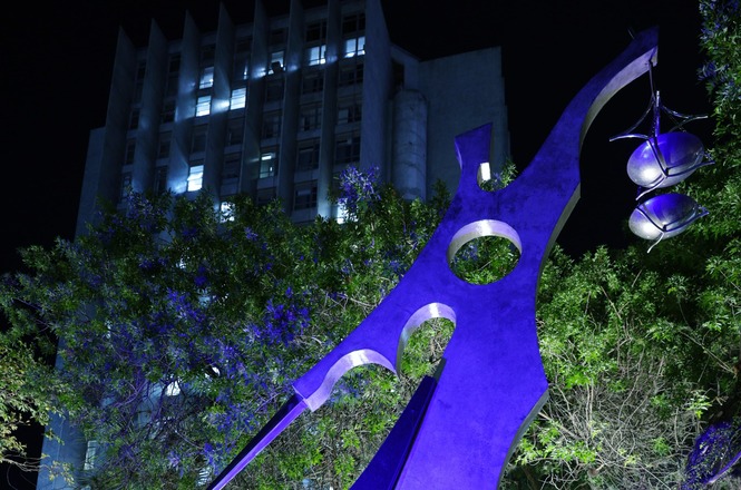 A imagem mostra uma escultura moderna grande, iluminada por luzes roxas, com uma forma abstrata estilizada. Ao fundo, há um edifício alto com janelas iluminadas contra o céu noturno. Árvores com folhagem verde cercam a área, adicionando um contraste natural à cena urbana.