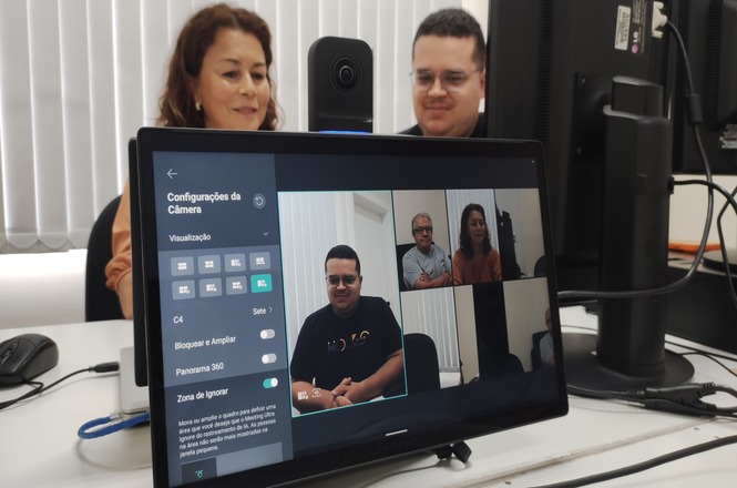 A imagem mostra duas pessoas sentadas em frente a um monitor de computador. O monitor exibe uma interface de configurações de câmera para videoconferência. Na tela, há janelas mostrando imagens de pessoas em uma videochamada. Há também uma câmera posicionada acima do monitor, voltada para os participantes da reunião. Além disso, há cabos conectados à mesa e equipamentos de informática ao redor.