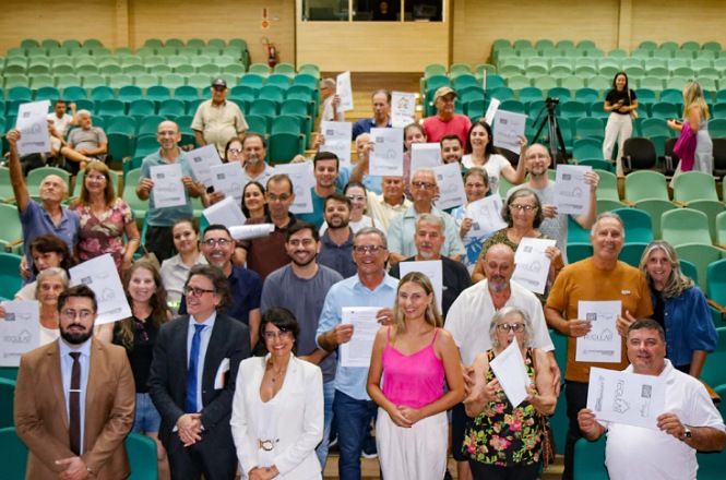 A imagem mostra um grupo de pessoas em um auditório posando para uma foto coletiva. Muitas delas estão segurando certificados ou documentos, aparentando ser uma cerimônia de formatura ou entrega de certificados. O auditório possui várias cadeiras verdes e algumas pessoas estão vestidas de forma formal. O ambiente parece ser alegre, com pessoas sorrindo para a câmera.