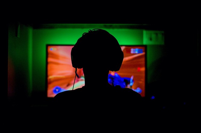 A imagem mostra a silhueta de uma pessoa usando fones de ouvido em frente a uma tela de computador. A tela está iluminada com cores vibrantes, principalmente laranja e azul, sugerindo que a pessoa está jogando um videogame. O ambiente ao redor está escuro, destacando ainda mais a luz da tela.