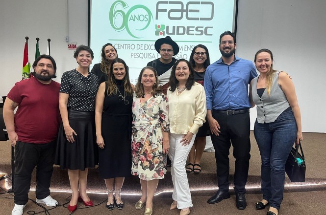 A imagem mostra um grupo de onze pessoas posando para uma foto em frente a um projetor que exibe um slide. O slide tem o logotipo "FAED UDESC" e menciona "60 anos". As pessoas estão bem vestidas, algumas com roupas formais e outras casuais. Há bandeiras no canto esquerdo da imagem, indicando que o evento pode ter um contexto institucional ou oficial. Todos estão sorrindo para a foto.