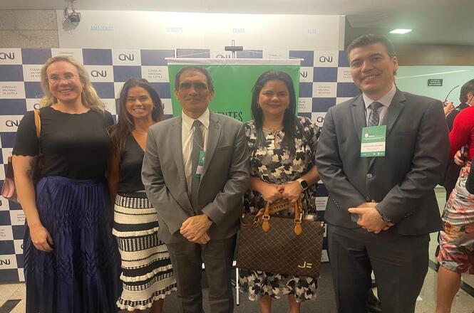 A imagem mostra um grupo de cinco pessoas posando para a foto em um evento. Eles estão de pé em frente a um fundo com logotipos do CNJ. Duas das pessoas estão vestidas formalmente com ternos, enquanto as outras três estão com roupas sociais. Eles estão sorrindo e parecem estar em um ambiente interno, possivelmente em um tipo de conferência ou reunião. Uma das pessoas está segurando uma bolsa.