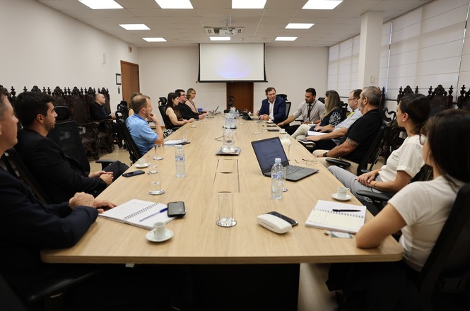 A imagem mostra uma sala de conferências onde várias pessoas estão reunidas ao redor de uma mesa retangular de madeira. Há papéis, laptops e garrafas de água sobre a mesa. As pessoas estão sentadas em cadeiras, parecendo participar de uma reunião ou discussão. No fundo, há uma tela grande, possivelmente para projeção. As pessoas parecem atentas, algumas tomando notas. A sala tem iluminação de teto e janelas com persianas fechadas.
