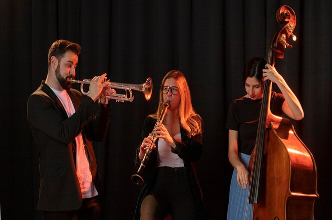 Na imagem, há três pessoas tocando instrumentos musicais. A pessoa à esquerda está tocando um trompete, a pessoa ao centro está tocando uma clarineta, e a pessoa à direita está tocando um contrabaixo. Todos parecem estar concentrados em sua performance musical. O fundo é escuro, destacando ainda mais os músicos e seus instrumentos.
