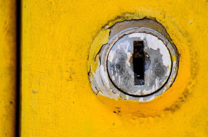 A imagem mostra uma fechadura de porta cercada por uma superfície amarela desgastada. A pintura ao redor da fechadura está descascada, revelando partes do metal por baixo. A fechadura é prateada, com sinais de uso e desgaste visíveis.