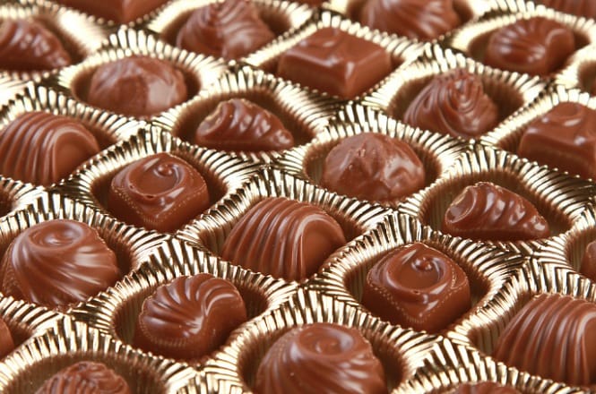 A imagem mostra uma caixa com diversos bombons de chocolate. Cada bombom está em um espaço individual e possui diferentes formas e designs na superfície. A embalagem é dourada, o que destaca os chocolates.