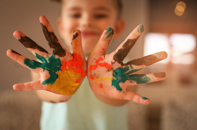 Na imagem, há uma criança desfocada ao fundo, estendendo as mãos para a câmera. As palmas de suas mãos estão cobertas de tinta colorida, incluindo cores como marrom, amarelo, laranja, verde e azul. A imagem transmite uma sensação de diversão e criatividade.
