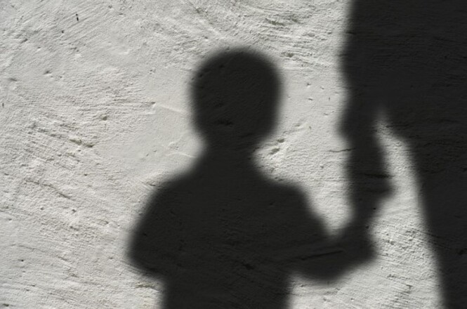 A imagem mostra a sombra de uma criança segurando a mão de um adulto em uma superfície clara. O contorno da criança está claramente definido, destacando a figura das duas pessoas projetadas na parede. A cena transmite uma sensação de proteção e cuidado.
