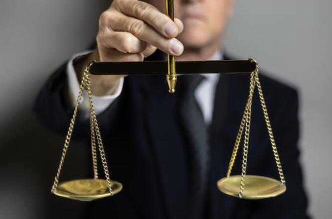 A imagem mostra uma pessoa segurando uma balança dourada. A balança é um símbolo tradicional da justiça, representando equilíbrio e equidade. A pessoa está vestida de forma formal, usando um terno escuro, mas o foco principal da imagem é a balança que está sendo segurada.