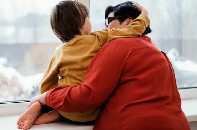 A imagem mostra um adulto e uma criança sentados em uma janela. A criança está vestindo um suéter amarelo e abraça o adulto de cabelos escuros e óculos, que veste uma camisa vermelha. Eles estão de costas para a câmera, olhando para fora. A cena parece ser de um momento carinhoso e tranquilo.