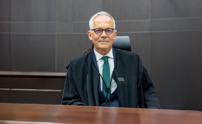 A imagem mostra um homem de meia-idade sentado em um ambiente que parece ser uma sala de tribunal. Ele está usando um terno escuro, gravata verde e uma toga preta, sugerindo que ele pode ser um advogado ou juiz. O fundo é de madeira escura, combinando com a mesa em frente a ele. Ele usa óculos e exibe uma expressão neutra.