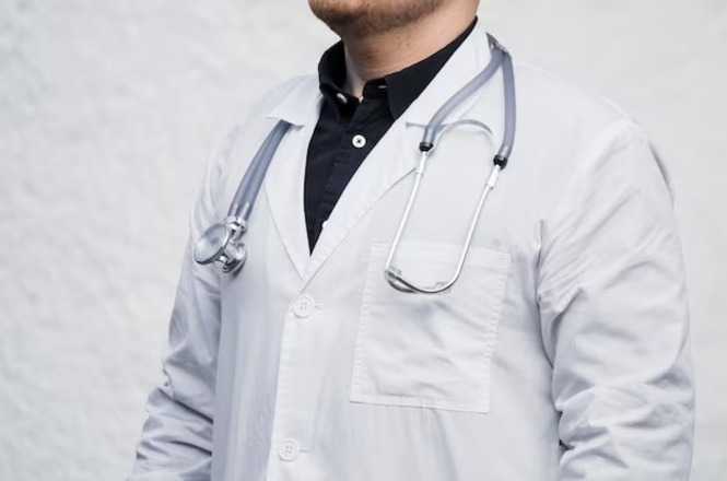 A imagem mostra uma pessoa vestindo um jaleco branco, que é típico de profissionais da área da saúde, como médicos. A pessoa está usando um estetoscópio em volta do pescoço. A camisa preta sob o jaleco contrasta com a vestimenta branca. O fundo é neutro, ajudando a destacar a figura principal na imagem.