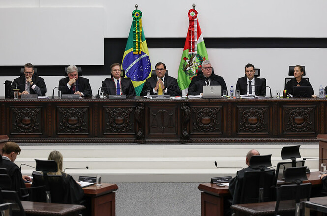 Na imagem, há um grupo de pessoas em um ambiente formal, possivelmente um tribunal. Elas estão sentadas atrás de uma bancada de madeira elaborada. No fundo, há duas bandeiras: a bandeira do Brasil e a bandeira de um estado brasileiro. As pessoas vestem trajes formais, sugerindo possivelmente magistrados ou advogados em uma sessão judicial. O cenário é de um julgamento ou deliberação oficial.