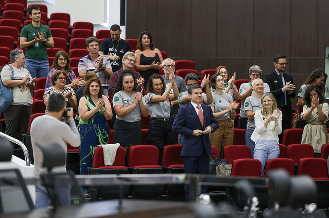 Na imagem, várias pessoas estão reunidas em um auditório. Elas estão de pé, aplaudindo, e algumas pessoas parecem estar usando uniformes ou camisetas semelhantes. O auditório tem cadeiras estofadas vermelhas. As expressões são de atenção e satisfação.
